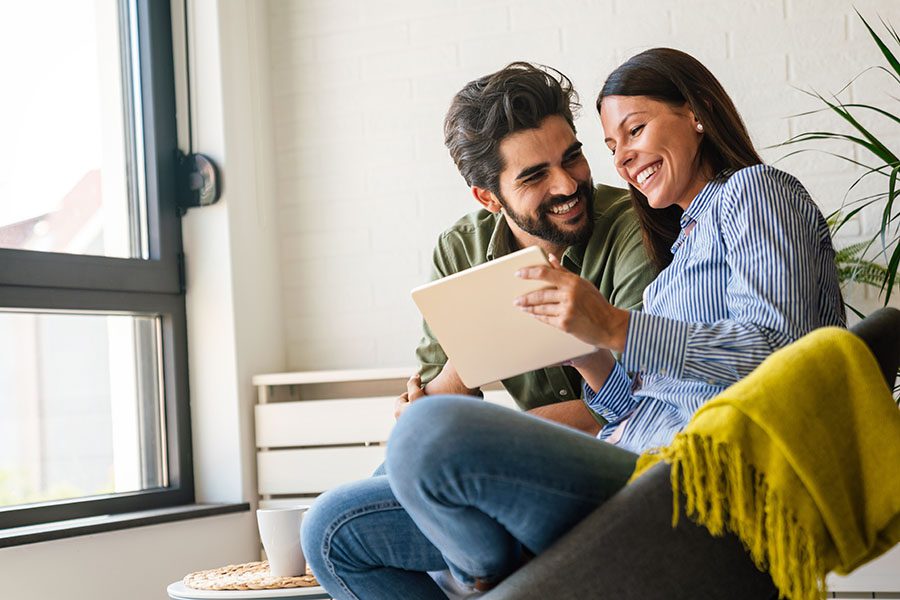 About Our Agency - Portrait of a Cheerful Young Couple Sitting in Their Living Room Having Fun Using a Tablet Together